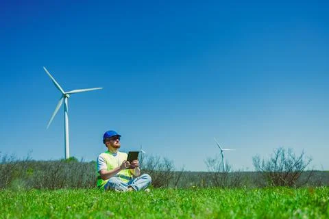 A windmill engineer sits on the grass holding a tablet while working near win Stock Photos