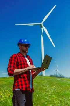 A windmill engineer stands on grass with a laptop, focused on work. Large win Stock Photos