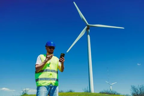 Windmill engineer stands on grassy field holding smartphone. Nearby is a larg Stock Photos