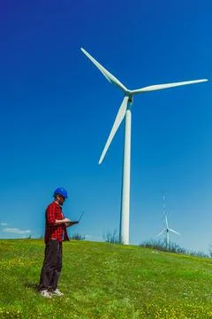 A windmill engineer stands on green grass, using a laptop while observing a t Photos