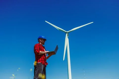 Windmill engineer stands under a clear blue sky using a laptop to check data  Stock Photos