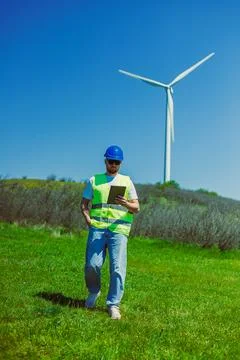 A windmill engineer walks through a green field holding a tablet. In the back Stock Photos