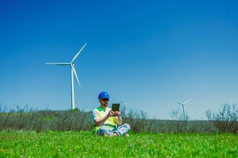 Windmill engineer working on a tablet in a grassy field under a clear blue .. Stock Photos