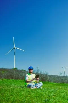 A windmill engineer works on a project while seated on grass near wind turbin Foto stock