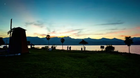 A windmill stands by a lake at sunset with mountains in distance Stock-Footage 312303902