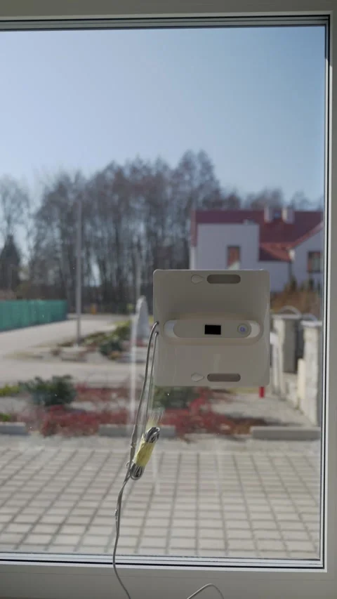 Window cleaning robot with hanging cord and residential yard visible outside Stock Footage 330880668