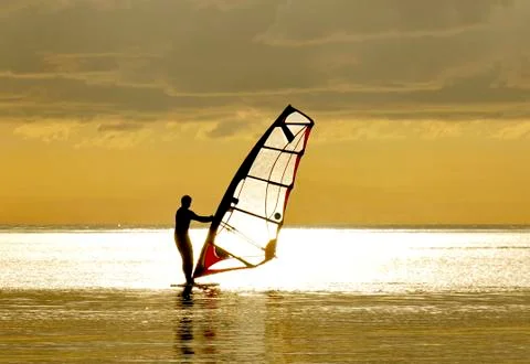 Windsurfing in the rays of the setting sun. Silhouette of a man on a golden b Stock Photos