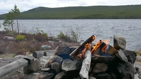 Windy lakeside scene with campfire burning among rocks, smoke drifting in wind Stock Footage 318196956