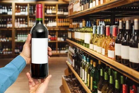 Wine wine bottle with empty label in female's hand in front of liquor store. Stock Photos