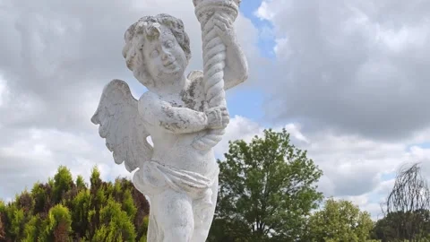 Winged marble statue outdoors gripping twisted rope above garden greenery Video stock 343628560