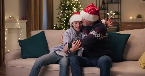 Winter evening. A Christmas tree flickers in room. A smiling young man hugs 스톡 동영상 210604131