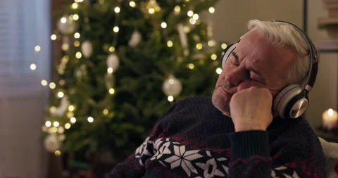 Winter evening. A senior man sits by Christmas tree and listens to relaxing 스톡 동영상 210580139