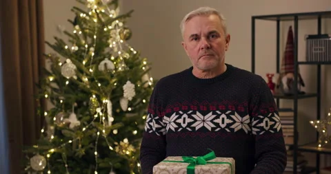 Winter evening. An upset senior man is standing by Christmas tree holding gift Stockbeeldmateriaal 210587757