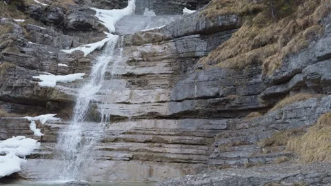 Winter waterfall flowing over layered rocky cliff with patches of ice and snow. Stock Footage 333223278