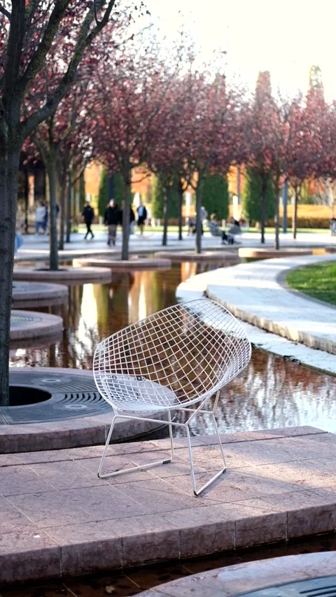 Wire chair positioned beside tranquil waterway with blooming trees in park Video stock 325486109