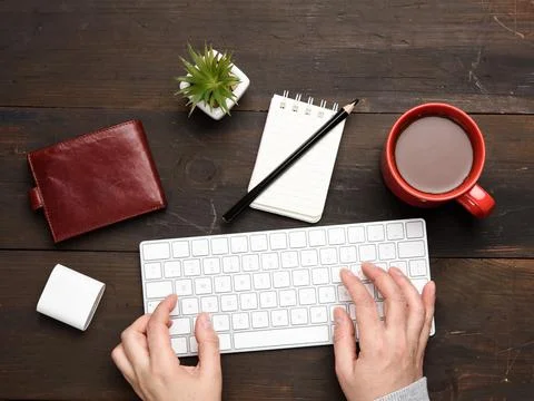 Wireless keyboard and two hands are typing on a wooden table, next to a cup w Stock Photos