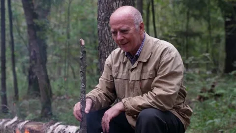 A wise thoughtful old man is sitting alone by a campfire in the forest, spending 스톡 동영상 166078083