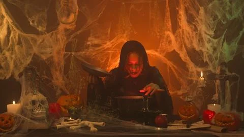 A witch in black clothes is cooking a potion in a scary kitchen with pumpkins Stock Photos