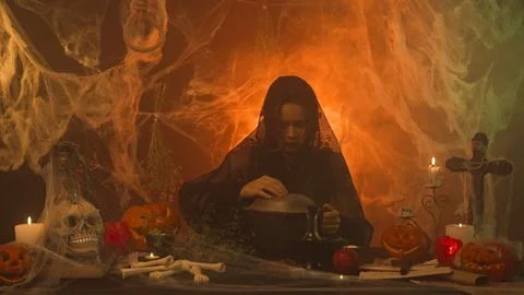 A witch in black clothes is cooking a potion in a scary kitchen with pumpkins Stock Photos