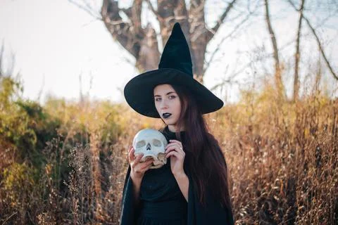 Witch with pale skin and black lips in a black hat, a dress and a raincoat Stock Photos