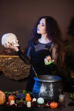 Witch with a skull in hands standing next to a cauldron and a table with herbs Stock Photos