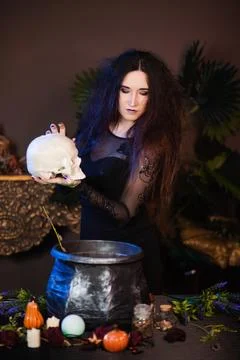 Witch with a skull in hands standing next to a cauldron and a table with herbs Stock Photos