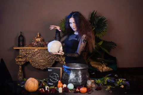 Witch with a skull in hands standing next to a cauldron and a table with herbs Stock Photos