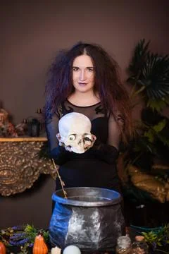 Witch with a skull in hands standing next to a cauldron and a table with herbs Stock Photos