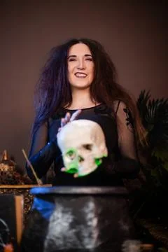 Witch with a skull in hands standing next to a cauldron and a table with herbs Stock Photos