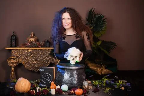 Witch with a skull in hands standing next to a cauldron and a table with herbs Stock Photos