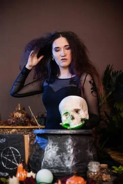 Witch with a skull in hands standing next to a cauldron and a table with herbs Stock Photos