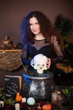 Witch with a skull in hands standing next to a cauldron and a table with herbs Stock Photos