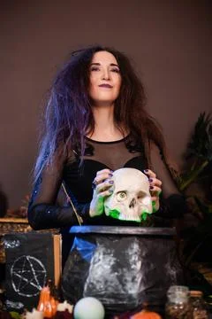 Witch with a skull in hands standing next to a cauldron and a table with herbs Stock Photos