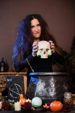 Witch with a skull in hands standing next to a cauldron and a table with herbs Stock Photos