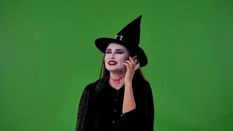 A witch with vampire makeup, in black cloak and hat, talks on her smartphone and Video stock 316587679