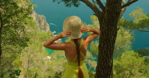 Woman adjusts her hat while viewing the cliffs and blue sea near Faraglioni in Stock-Footage 327612714