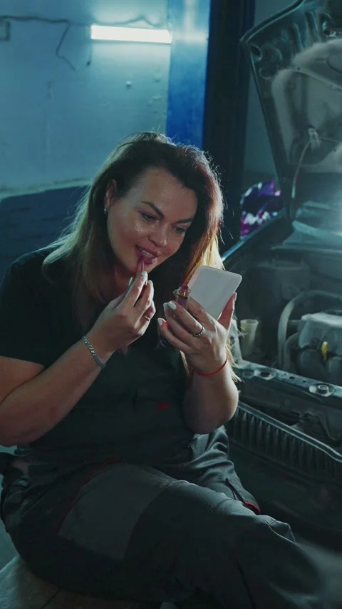 Woman applies makeup while sitting beside a car engine in a garage Stock Footage 303797891