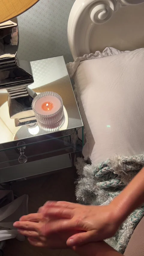 Woman applying hand cream next to bed and lit candle in cozy bedroom Video stock 346094853