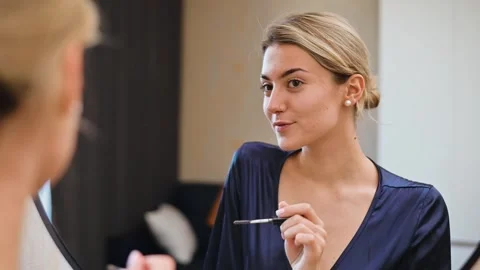 A woman is applying makeup while seated in a well-lit room, using various Stock Footage 337555156