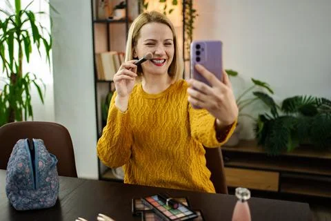 Woman applying makeup while taking a selfie in a cozy room with plants and Stock Photos