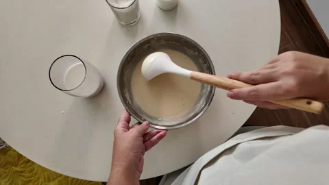 A woman in an apron, using a ladle, is stirring pancake batter in a metal bowl Stock Footage 319345443