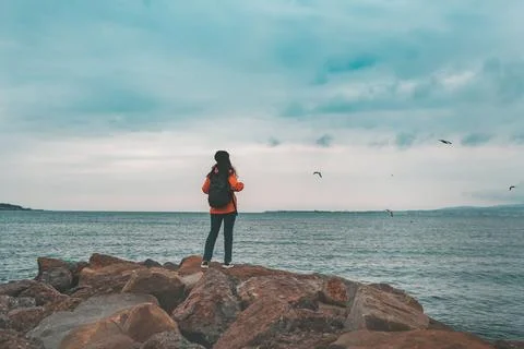 A woman with a backpack in an orange jacket and hat stands on the coastal bou Stock Photos