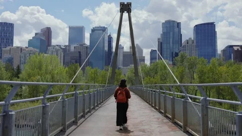 Woman with backpack walking on pathway bridge with Calgary city skyline in 스톡 동영상 330952845