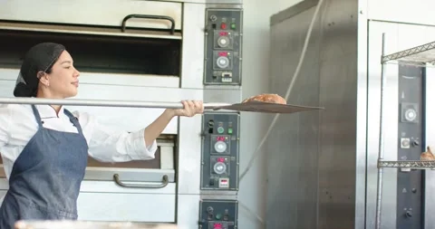 Woman baker hearing oven timer, removing loaves with peel onto cooling shelves Stock Footage 343517710