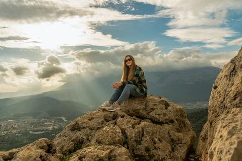 A woman basks in the sunlight while sitting on a high mountain, with a city at t Stock Photos