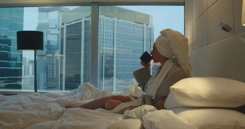 Woman in bathrobe drinking coffee on bed in luxury hotel city view Stock Footage 334426547