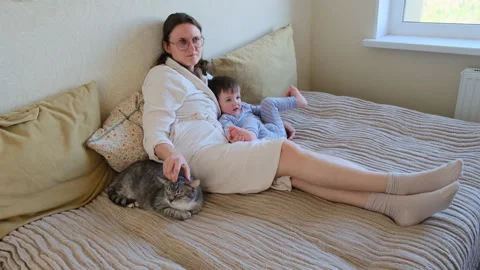 Woman in bathrobe sitting on bed with small child and tabby cat. Relaxed mother Stock Footage 345980698