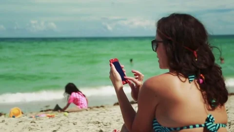 Woman at beach using cell phone while watching daughter by ocean Stock Footage 78385532