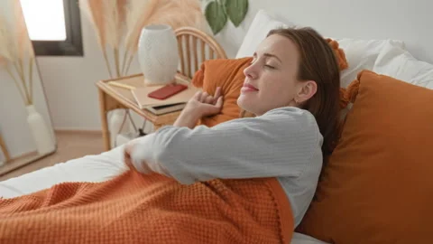 Woman on bed pulls orange knit blanket and stretches arm toward window in c.. Stock Footage 337434600