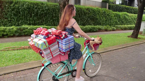 Woman biking with large stack of gift boxes on rear rack, cute dog in basket Stock Footage 301001752